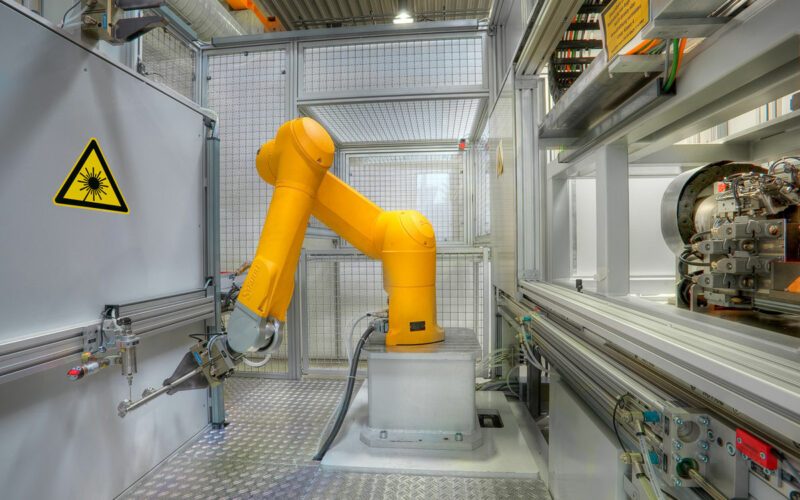 Laserheften, Maschine von innen, gelber Roboterarm in der Mitte bewegt sich