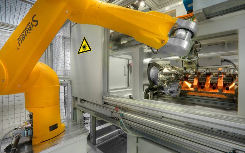 Laserheften, Maschine von innen, gelber Roboterarm in der Mitte bewegt sich und fügt etwas seitlich in die Maschine ein