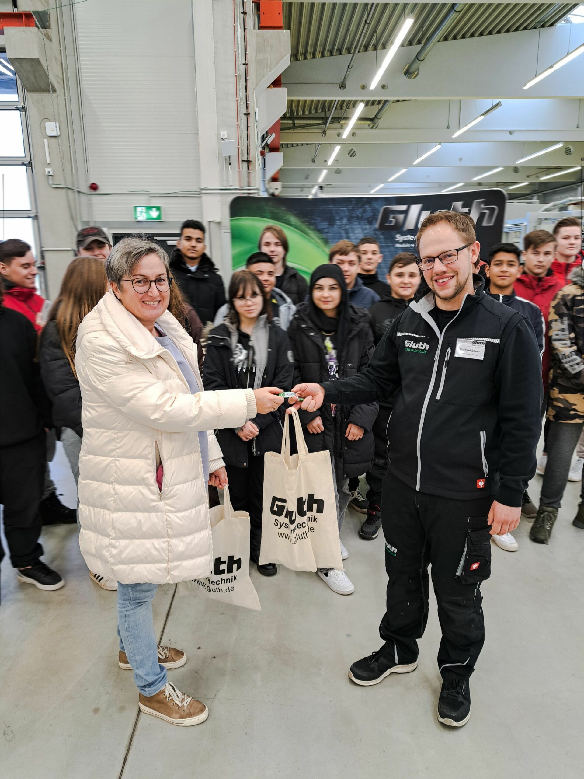 Mittelschule St. Stephan zu Besuch bei Gluth, eine Lehrerin reicht einem Mitarbeiter der Firma Gluth etwas, beide lächeln in die Kamera